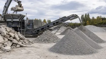 En grävmaskin matar betong i en kross intill högar med krossmaterial.